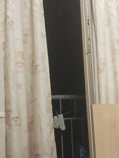 This media file shows a glimpse through an open window at night. The scene is dimly lit, with the predominant colors being muted beiges and grays. On the left, a patterned curtain with a floral or abstract design hangs, partially obscuring the view. To the right of the curtain, the dark interior of a room is visible, with a window frame and latch clearly depicted. Beyond the window, a dark space is seen, possibly an outdoor area or another room. There is a thin horizontal line, possibly a clothesline, from which a small, light-colored object, appearing to be a piece of clothing, hangs. Behind this, a metal railing is visible, suggesting a balcony or external structure. The lighting indicates it is nighttime, and there are no specific weather cues. The overall impression is one of a domestic interior, possibly a bedroom or living area, with a view outside. No text is visible in the image. The setting appears to be a residential building.