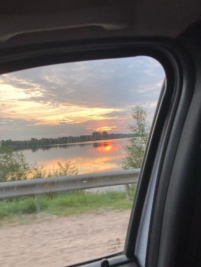 The image depicts a view from inside a vehicle, looking out through a car window. The scene outside is a landscape at sunset, with the sun low on the horizon, casting a warm glow across the sky and reflecting on a body of water. The sky is filled with dramatic clouds, tinged with orange, yellow, and pink hues. A line of trees is visible along the edge of the water. On the right side of the image, there is a dirt or gravel road and some green foliage, possibly grass or trees, partially obscured by a reflective barrier or part of the car's exterior. The interior of the car is visible in the foreground, with dark, textured surfaces framing the window.
The time of day is clearly sunset, indicated by the low sun and the colors in the sky. The weather appears to be clear with scattered clouds. The location is hinted at by a small piece of text on the car window, which appears to be a vehicle registration or compliance sticker, and has Cyrillic characters, consistent with the specified location of Niasvizh, Belarus. No people are visible in the image. The overall mood is peaceful and serene, capturing the beauty of a sunset over a natural landscape.