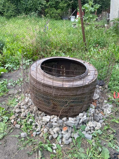 In the city of Lebyazhye, Russia, a scene unfolds in an outdoor, overgrown environment. The central focus is a repurposed washing machine drum, serving as a makeshift fire pit. This metallic cylinder, exhibiting signs of rust and age, is encircled by a cage of wire mesh. The drum rests on a bed of assorted grey and reddish stones and rubble. Surrounding the fire pit is a patch of uneven ground, sparsely covered with green grass and scattered weeds, indicating a neglected or rural setting. Beyond this immediate area, a chain-link fence runs horizontally, separating the foreground from a densely vegetated background of green bushes and trees. In the far distance, parts of structures and possibly wooden debris are visible, suggesting the presence of an industrial or storage area. The lighting suggests it is daytime, with the sun casting diffuse light on the scene. There are no people present in the image.