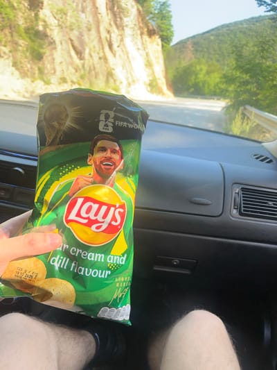 The image shows a hand holding a bag of Lay's sour cream and dill flavored chips in the passenger seat of a car. The bag features a picture of Lionel Messi and the FIFA World Cup logo. Outside the car, a winding road is visible, with a steep, rocky incline on the left and lush green trees on the right, indicating a mountainous or hilly area. The scene appears to be on a road near Taran, Bulgaria, characterized by its natural landscape.