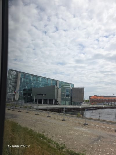 The image shows a modern glass-fronted building with a large "C" logo and the text "BUREAUX A LOUER" and a phone number visible. The building is located in the port of Le Havre, France. In front of the building, there is a body of water, a harbor, and a walkway with metal barriers. To the right of the main building, there is a red brick building. The sky is cloudy. A date stamp in Cyrillic script "15 юни 2026 г." is superimposed in the lower left corner.