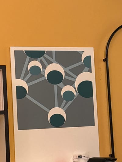 The image displays a piece of abstract art featuring a network of interconnected circles and lines against a dark gray background. The circles are divided into two colors: white on one side and a deep teal on the other. The overall composition is geometric and minimalist. The artwork is framed by a white border and appears to be leaning against a wall painted a warm, mustard yellow. To the left of the artwork, a portion of a black frame is visible, possibly belonging to another piece of art or a picture frame. On the right side, a thin, curved black metal rod is present, extending vertically. At the bottom right, a stack of white books or binders and a black wallet are placed on a surface. There is no indication of the city of Diegem, Belgium, or any specific location within it. The image is a close-up, cropped view, and no people are present in the scene.