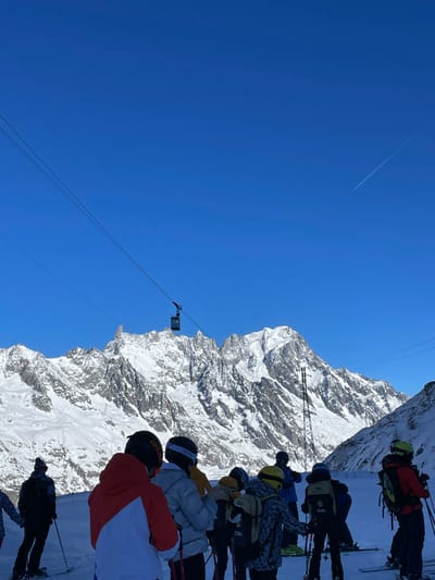 The image, taken on a bright, sunny day, depicts a group of skiers in a high alpine environment, likely Courmayeur, Italy. In the foreground, several individuals, dressed in full winter ski gear including helmets, goggles, and backpacks, stand on skis. Most are facing away from the camera, looking towards the vast, snow-covered mountain range that dominates the right side of the frame. Visible brands on their equipment include "SMITH" and "SALOMON" on helmets, and "CVA" on multiple backpacks.

The sky on the left is a clear, deep blue, with a single white airplane contrail visible in the upper left quadrant. In the mid-ground, a cable car cabin is suspended by cables against the backdrop of the mountains, suggesting a ski lift system. A tall metal structure, possibly a support pylon for the lift, also rises from the snowy slopes. The mountains are rugged, characterized by dramatic rocky outcrops and expansive snowfields, reflecting the bright sunlight.

The scene captures a moment of anticipation or gathering, possibly before a descent or while waiting for a lift. The clear weather and pristine snow suggest ideal conditions for skiing or snowboarding, amidst a breathtaking natural landscape.