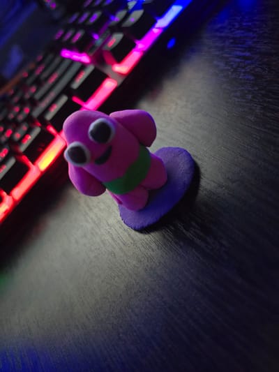 A small, pink, clay figure with large black and white eyes and a green band around its torso is positioned on a purple heart-shaped base. The figure is placed on a dark, textured surface, possibly a wooden table. In the background, a keyboard with illuminated keys in red, pink, and blue is visible, suggesting a gaming or computer-related setting. The scene is dimly lit, with the keyboard's lights providing the primary illumination. There are no people or any identifiable landmarks to suggest a specific location in the city of Shali, Russia. The image is taken from a low angle, with a shallow depth of field that blurs the background.