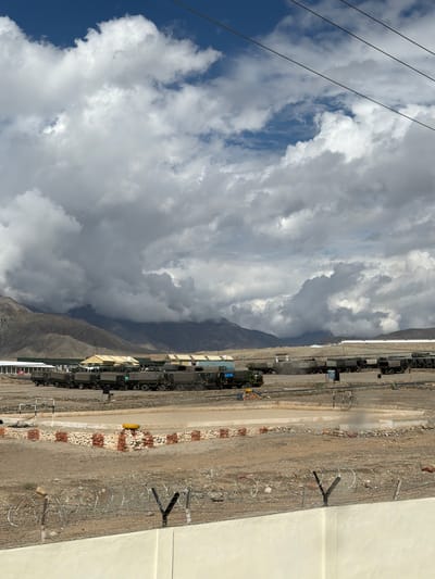 The image shows a military base with several trucks lined up. The base is located in a mountainous area with a cloudy sky. There are also some buildings and a fence in the foreground. The scene appears to be a daytime outdoor shot. The trucks are of various sizes and colors, and they are parked in rows. Some of the trucks are covered with tarps. The buildings are made of concrete and have corrugated metal roofs. The fence is made of barbed wire and posts. The mountains in the background are covered with snow. The sky is filled with dark, heavy clouds, suggesting a stormy weather condition.