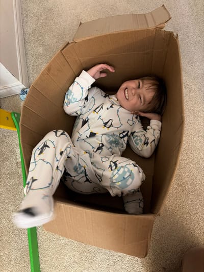 The image depicts an indoor scene within the city of Woodstock, Canada.

A single individual, a young child, is positioned lying on their back inside a large, open, brown cardboard box. The child is wearing a white full-body pajama suit patterned with black penguins, multi-colored string lights, and light blue Christmas ornaments. The child's head is slightly tilted to their right, and their arms are bent with hands near the head and chest. Both legs are bent at the knees. The child is looking directly towards the camera with an open-mouthed smile, revealing teeth, and eyes wide open.

The cardboard box is rectangular, with its top flaps splayed outwards. The interior of the box is empty save for the child. The box rests on a light-colored, short-pile carpeted floor. In the background to the left of the box, a portion of a child's toy or furniture piece, consisting of a bright green vertical element and a yellow component, is visible. A white baseboard and a small piece of blue fabric are also partially visible against a wall in the background. The child's left foot appears slightly blurred, indicating possible motion.