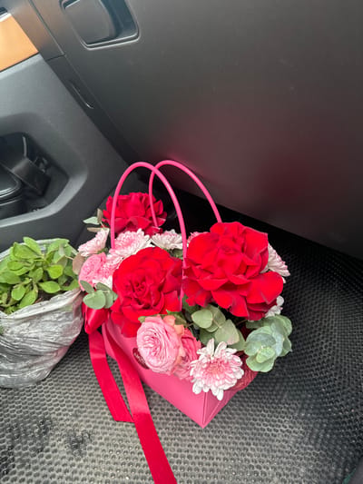A close-up, eye-level shot captures a bright pink flower arrangement, featuring vibrant red roses and delicate pink roses, nestled within a pink box with matching handles. The arrangement is adorned with a bright red ribbon. To the left of the flower arrangement, a small potted plant in a textured, gray wrapper is visible. The scene appears to be inside a vehicle, with the black dashboard and textured floor mat clearly visible. The background suggests the interior of a car, with a portion of a tan dashboard and a black seat visible on the left. The lighting is moderate, highlighting the colors of the flowers. The image does not contain any information about the location in the city of Сароцкі сельскі Савет, Belarus.
