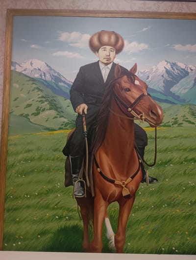 A painted portrait depicts a man on horseback. The man, wearing a dark suit, white shirt, and a large, round fur hat, has a mustache and a neutral expression. He holds the reins and a whip. The horse is brown with a white marking on its forehead and a white sock on its hind leg. The background shows a vibrant green field with scattered wildflowers, rolling hills, and snow-capped mountains under a blue sky with white clouds. The painting is framed by a golden border. The scene is set in a pastoral, mountainous landscape, likely representative of the natural beauty of Kyrgyzstan.