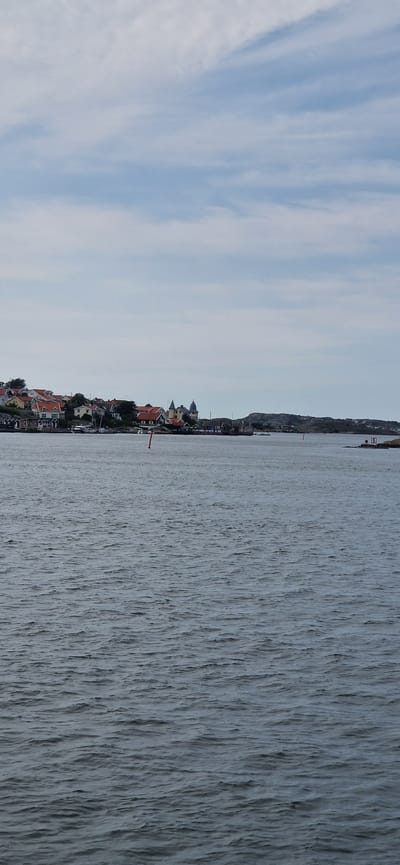 A calm body of water stretches across the lower half of the frame, with gentle ripples reflecting the overcast sky. On the horizon, a cluster of colorful houses with red roofs forms a picturesque village nestled along a rocky coastline. The village appears to be a settlement on Öckerö island, part of the Öckerö Municipality in Sweden, characterized by its traditional Nordic architecture and coastal setting. The sky above is a pale blue with streaks of white clouds, suggesting a cool, perhaps breezy, day. There are no people visible in the image, and the scene conveys a sense of peaceful, coastal life. A red buoy stands upright in the water, marking a channel or hazard, and a white marker with the number '5' is visible on a distant rocky outcrop.
