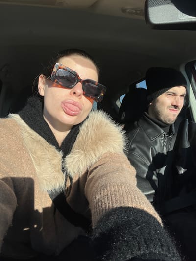 The scene takes place inside a vehicle in Marquette-lez-Lille, France, illuminated by strong, direct sunlight. A female passenger, positioned on the left, faces the camera. She wears large, tortoise-shell framed sunglasses, a light brown knitted garment with a light-colored faux fur collar, and a black garment underneath. Her mouth is open with her tongue slightly extended. To her right, in the driver's seat, a male individual is present. He wears a black knitted beanie, a black leather-like jacket over a black turtleneck, and has facial hair. His gaze is directed forward and slightly to the right, with a squinted or grimacing expression. The sun creates distinct shadows across the subjects and the gray interior of the vehicle, highlighting the passenger compartment. A rear-view mirror is visible above and between the individuals.