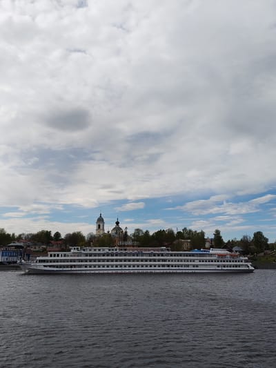 A long, white passenger cruise ship is on the water in front of Myshkin, Russia. The ship has multiple decks and appears to be docked or moving very slowly along the river. The skyline of Myshkin is visible in the background, with a church featuring a tall, light-colored bell tower and dark-colored domes standing out amidst the trees and other buildings. The sky is filled with a mix of white and grey clouds. The water is dark, with slight ripples on the surface.