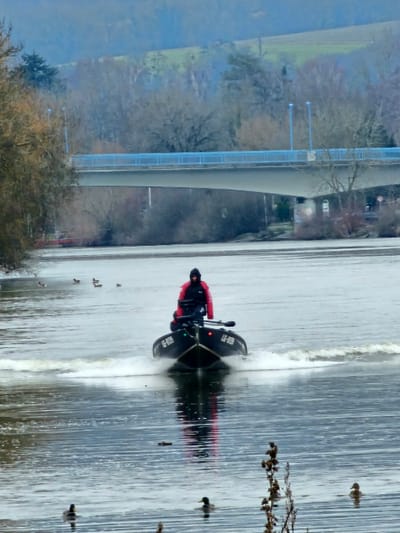 A dark-colored rigid inflatable boat (RIB) displaying the identification 'LG-R039' is observed moving on the Moselle River near Bech-Kleinmacher, Luxembourg, generating a wake. A lone individual, wearing a red and black jacket with a dark hood, operates the boat from the stern. The boat is equipped with forward-facing navigation lights and an oar stowed on its port side. Multiple ducks are scattered on the water surface in the foreground and mid-ground. In the background, the Hëttermillen Bridge (B12), a concrete structure with light blue railings, spans the river. The banks beneath the bridge are vegetated with bare deciduous trees and shrubs. Beyond the river, rolling hills covered with a mix of leafless forests and green fields extend into the distance under an overcast sky.