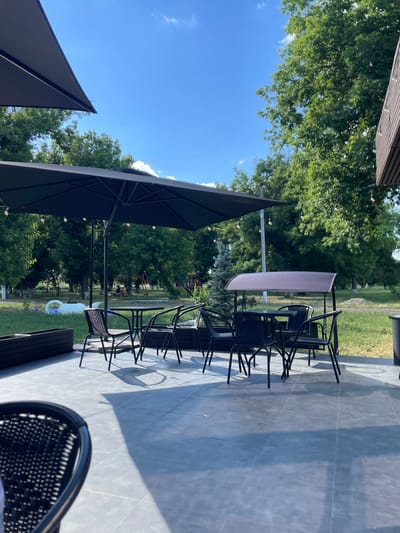 The image depicts an outdoor seating area of a cafe or restaurant on a sunny day. Several black metal tables and chairs are arranged on a tiled patio. Two large black umbrellas provide shade over some of the tables. Beyond the patio, there is a grassy area with trees and a playground in the background. The location is identified as Znamianka, Ukraine. There are no people visible in the immediate seating area, but some figures can be seen in the distance on the playground. The environment appears to be a park or public space adjacent to the establishment.