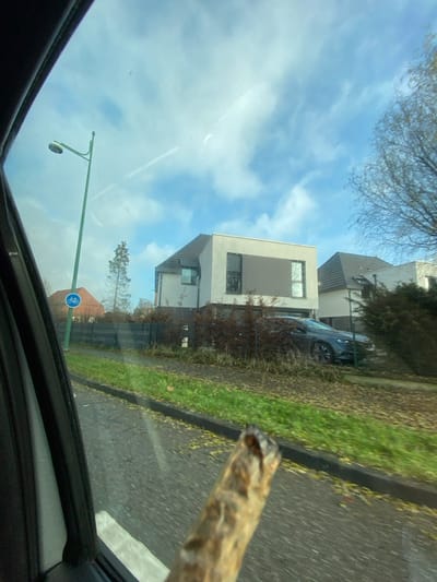 The image depicts a street scene in La Wantzenau, France, captured from the interior of a vehicle looking out the front passenger side window. In the immediate foreground, a blurred, partially consumed, hand-rolled item with a lit end is held protruding from the vehicle's window frame.
The midground features a paved road surface alongside a curb, bordered by a strip of green grass interspersed with scattered brown leaves. A tall, green lamppost with a circular blue sign depicting a white bicycle is positioned to the left, indicating a cycle path.
Beyond the grassy strip, a dark horizontal fence runs in front of modern residential properties. The background includes several houses, prominently a two-story residence with a light-colored facade, dark-framed large rectangular windows, and a dark-tiled, sloped roof. A dark-colored sedan is parked in the driveway or on the property directly in front of this house. Various trees are visible, with some deciduous trees appearing bare, suggesting late autumn or early winter. The sky is blue with scattered white clouds and at least two visible airplane contrails. The scene is illuminated by daylight.