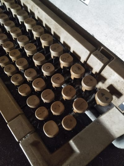 The image displays a close-up, angled view of the keyboard of an aged mechanical typewriter, located in Жібек Жолы, Kazakhstan. The keyboard features a QWERTY layout with multiple rows of circular, light brown/beige keys bearing dark imprinted characters, including letters (e.g., 'Q', 'W', 'E', 'R', 'T', 'Y', 'U', 'I', 'O', 'P', 'S', 'D', 'F', 'G', 'H', 'J', 'K', 'L', 'N', 'M'), numbers (e.g., '2' through '0'), and symbols (e.g., '/', '.', '+', '-', '1/4', '1/2'). A rectangular "TAB CLEAR" key is visible on the upper right section of the keyboard. The typewriter's frame, also light brown/beige, surrounds the keys. A dark brown space bar is partially visible at the bottom. The surfaces of the keys and the frame show noticeable wear, accumulation of dust, and small particulate matter. The background consists of a light-colored surface with a small, indistinct white object in the upper right. The scene is illuminated, casting minor shadows and highlighting surface textures. No individuals or active processes are depicted.