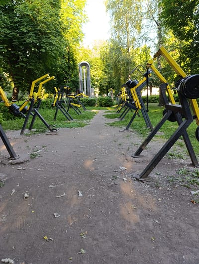 This media file depicts an outdoor exercise area with a row of weight machines. The machines are primarily yellow and black, with visible weight plates on some. They are arranged along a dirt path bordered by grass and trees. In the background, a tall, modern-looking archway structure is visible, suggesting a park or public space.
The scene appears to be set during the day, with sunlight filtering through the leaves of the trees, casting dappled shadows on the ground. The foliage suggests a temperate climate, likely during spring or summer. The overall mood is one of tranquility and a focus on physical activity. There are no people visible in the image, and no visible text. The location context provided is Fastiv, Ukraine.