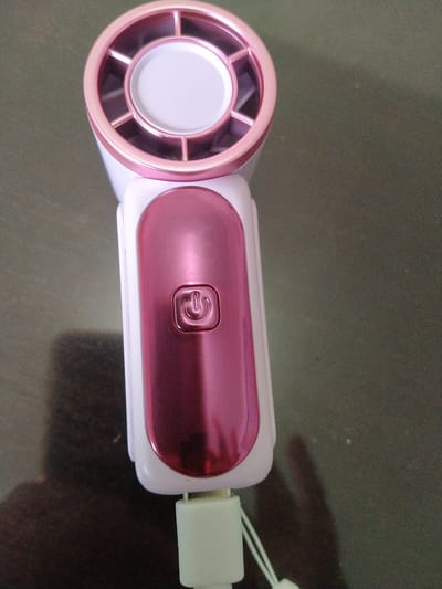 A handheld pink and white fan with a metallic pink oval power button is shown from a slightly elevated angle, lying on a dark, textured surface. The fan has a circular opening at the top, surrounded by a rose gold-colored rim, with white internal blades. The power button, located on the main body of the fan, is also metallic pink and features a circular power symbol. A white USB cable is attached to the bottom of the fan, with a white plastic connector. The background is a dark, speckled surface, likely a tabletop. There are no people visible in the image, nor any specific identifiable features that would indicate its location within Pulorayeuk, Indonesia.