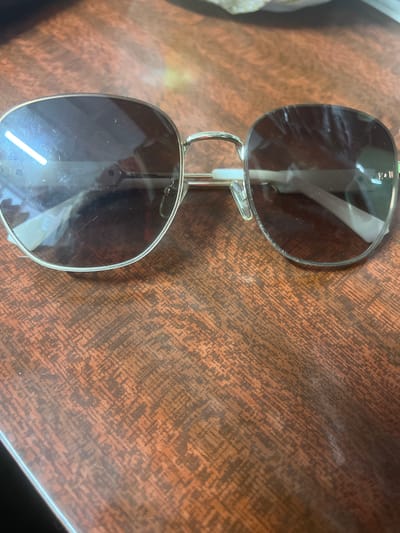 A pair of aviator sunglasses with silver rims and dark lenses rests on a brown, textured wooden surface, possibly a table. The sunglasses are positioned slightly open, with the left lens facing forward and slightly angled towards the viewer. The surface appears to have a wood grain pattern with variations in color, ranging from light brown to darker reddish-brown tones. There are some reflections visible on the lenses of the sunglasses, indicating light sources in the environment. The image is taken from a high angle, looking down at the sunglasses. The location in the city of Şū, Kazakhstan is not discernible from the provided image.