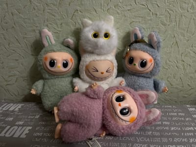 Four plush toys are arranged on a grey fabric surface with a repeating "LOVE YOU" pattern. The toys are designed to resemble fantastical creatures with large, round eyes, painted rosy cheeks, and wide smiles with small teeth. From left to right, the first toy is green with bunny ears. Next to it is a white toy with large yellow eyes and cat-like ears. To the right of the white toy is a blueish-grey toy with bunny ears and a small gold pendant or tag attached to its hood. In the foreground, lying on its back, is a pink toy with bunny ears, its body curved and its face looking upwards. The background consists of a textured wall with a subtle pattern and some glittering elements. The lighting appears to be artificial, casting soft shadows. The image is a still life, showing the toys in a static arrangement. The toys are clean and appear to be in good condition. There is no indication of the city of Muravlenko, Russia, in the image itself.