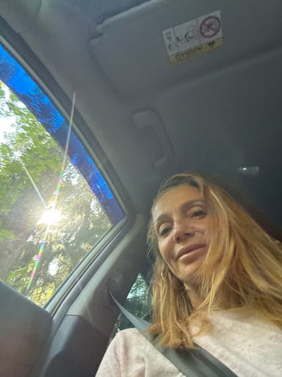A woman with blonde hair and a pink shirt is in the passenger seat of a car. She is looking at the camera. The sun is shining through the window, casting a rainbow effect on the car's interior. The car is in the city of Portovoe, Russia.