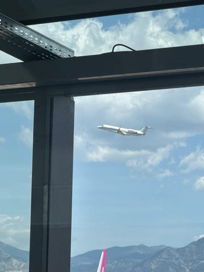 An aerial view from a building, showing a white airplane flying in a cloudy sky. The plane is viewed from the side and slightly from below, with its wings extended. The foreground shows a dark, modern building structure with glass windows. In the background, mountains are visible under a bright sky with scattered clouds. A small portion of a pink airplane tail fin is visible at the bottom of the image. This scene is located in Kruja, Albania.