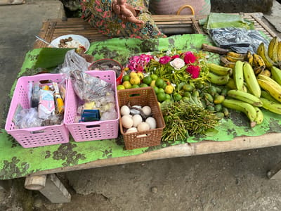 This image captures an outdoor market scene in Bumireja, Indonesia. A table made of bamboo and wood displays various items for sale, including a plate of rice and side dishes, pink plastic baskets filled with packaged goods and what appear to be cigarettes, a brown basket containing a dozen eggs, a pile of green fruits (likely limes or small oranges), bunches of green produce with long stems, and several bunches of green bananas and yellowing bananas. Flowers, including pink and white roses, are arranged on top of the green produce. A person, partially visible, is seated behind the table, wearing a colorful patterned garment and resting their hands on the table. The surface of the table is covered with a green cloth featuring a dark floral pattern. The ground beneath the table appears to be rough and uneven.