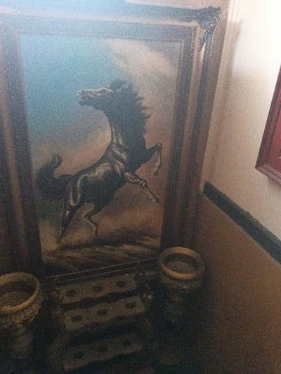 The image displays an indoor setting in Dogsenang Gorphoog, Bhutan, dominated by a large, vertically oriented painting of a black horse. The horse is depicted in a rearing posture, facing left with its head slightly turned, against a background that transitions from a light blue sky at the top to a darker, earthy tone at the bottom. The painting is encased in a dark, ornate frame, exhibiting a decorative element at its upper right corner. Below the painting, on a dark surface, are two light-colored, urn-shaped objects with circular openings, positioned on either side of a central, dark, multi-tiered stand. This stand presents at least three visible levels, each featuring multiple circular indentations. To the right of the painting, a light brown wall is visible, marked by a dark horizontal molding or rail. Above this molding, a smaller object with a dark red frame is partially visible on the wall. The scene is dimly lit and the image exhibits a grainy texture.
