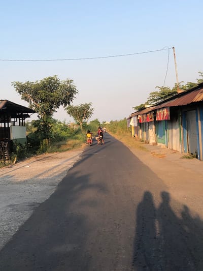 In Glagah Arum, Indonesia, a rural road scene unfolds under a clear sky. The road, paved and dark, dominates the foreground, with its surface showing some gravel and a distinct shadow cast by an unseen object, likely trees, creating a dramatic contrast. To the left of the road, a simple, open-sided structure with a corrugated metal roof stands, partially obscured by dense green foliage and trees. The vegetation is lush and extends to the right of the road as well, creating a natural border.
Further down the road, two figures are riding small motorcycles. The first motorcyclist, wearing a yellow shirt, is ahead. The second motorcyclist, in a black shirt, is following closely. The buildings lining the right side of the road are simple, constructed with a variety of materials, including brick, wood, and corrugated metal for the roofs. The structures appear to be shops or small dwellings, with colorful cloths or banners hanging from the overhangs. One building features blue doors or shutters. The overall atmosphere is that of a quiet, everyday scene in a village setting.