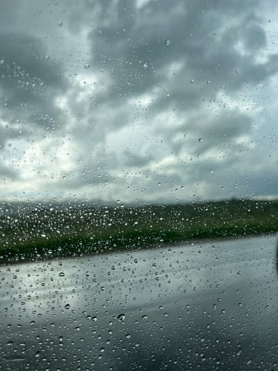 The image depicts a view through a rain-streaked window, likely from inside a vehicle, looking out at a cloudy, overcast sky and a landscape. The foreground is dominated by numerous water droplets of various sizes adhered to the glass surface, distorting the view of the scene beyond.
The environment visible through the window includes a dark, wet road surface in the lower right quadrant of the image. Above the road and to the left, there is a band of green vegetation, possibly rolling hills or fields, suggesting a rural or semi-rural setting. In the background, a hazy, muted landscape can be perceived, with hints of distant hills or mountains. The sky is filled with heavy, grey clouds, indicating a significant weather event, such as rain or a storm.
The image does not contain any people, animals, or specific man-made structures that would definitively place it within the city of Apnagyugh, Armenia. The scene is purely atmospheric and environmental, focusing on the visual effects of rain on a window and the muted landscape beyond.