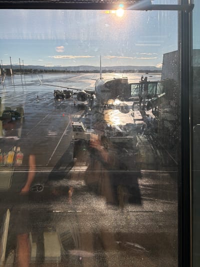 The image depicts an airport tarmac at Oslo Airport, Gardermoen, Norway. An airplane is connected to a jet bridge, with various ground support vehicles and equipment surrounding it. The tarmac appears wet, with reflections of the sky and buildings. Sunlight is casting a bright glare across the scene, with visible lens flare. In the foreground, the reflections on the window obscure some details, but suggest people inside the terminal looking out at the tarmac. The environment is overcast with some clouds and distant mountains visible.