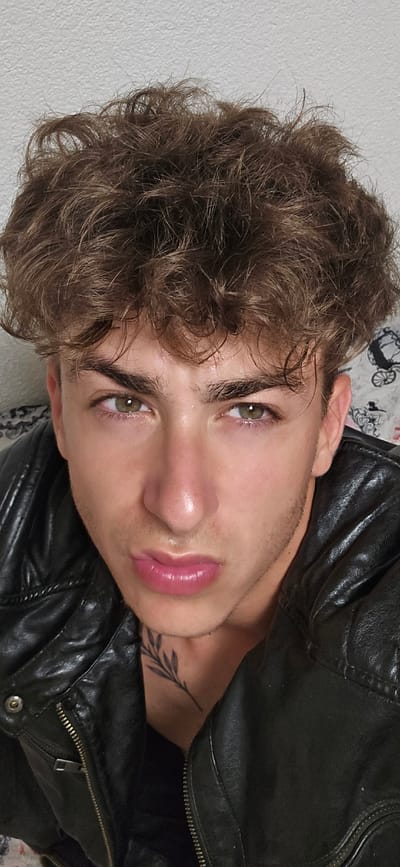 The image is a close-up, head-and-shoulders portrait of a young man with curly, brown hair and green eyes. He is wearing a black leather jacket. His lips are parted, and he appears to be looking directly at the camera. A small, black tattoo of a branch is visible on his neck. The background is a textured, light-colored surface, possibly a wall. There is no visible indication of the city of Kitzscher, Germany, or any other specific location.