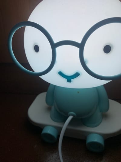 A close-up, slightly angled shot features a pale blue and white novelty night light in the shape of a character with large circular glasses and a gentle smile. The character is seated on a white base resembling a skateboard, complete with two teal-colored wheels visible at the bottom. A white power cord extends from the underside of the character. The background is a dark, textured surface, possibly wood. The scene is indoors, with no discernible elements suggesting a specific city or location beyond what is described.