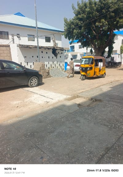 The scene depicts a street in Kofar Ruwa, Nigeria, under clear daylight conditions. The ground in the foreground is a mix of concrete paving, exhibiting visible wear and depressions, transitioning into an unpaved dirt and gravel area. A black sedan is parked on the left, partially within the frame. Adjacent to it, a stack of concrete blocks and a pile of gravel are present.
In the background, a two-story white building with a blue corrugated metal roof features multiple windows and an externally mounted air conditioning unit. The lower section of this building has a distinct white facade with numerous small, dark rectangular openings arranged in vertical and horizontal patterns, and an area of visible structural damage or unfinished construction. A slender, tall metal pole stands vertically in front of this building. To the right, a large tree with green foliage is prominent.
A yellow three-wheeled auto-rickshaw, marked "GMS" on its front, is positioned near the center-right, appearing stationary. One passenger is visible within the vehicle. A male individual, dressed in a light-colored, long-sleeved garment, walks from left to right past the auto-rickshaw, his back mostly facing the camera. Further in the background, a white sedan and a cluster of motorcycles are parked near a second white building, also topped with a blue roof. No direct interactions between individuals are discernible.