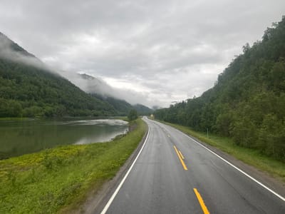 The image depicts a scenic view of a road winding through a mountainous landscape in Engan, Norway. The road is asphalt with distinct yellow and white lane markings, suggesting it is a well-maintained public highway. To the left of the road, a calm, wide river flows, its surface reflecting the overcast sky. The riverbank is lined with lush green grass and interspersed with patches of small yellow flowers. Dense, verdant forests cover the steep slopes of the mountains on both sides of the road. The mountains themselves are partially shrouded in mist, creating a dramatic and atmospheric effect. In the distance, a small, yellow house is visible nestled among the trees on the right side of the road. The sky is overcast with heavy, grey clouds, indicating a cloudy or possibly rainy day. There are no people or vehicles visible in the image.