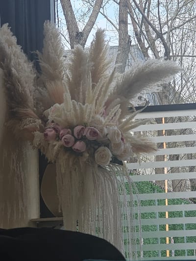The scene is an interior view overlooking an outdoor environment in Fundeni - Dobroești, Romania. The primary subject is a large, elaborate floral and botanical arrangement positioned in the mid-ground. This arrangement comprises numerous cream-colored pampas grass plumes, interspersed with clusters of artificial roses in shades of light pink and white. Below the main arrangement, several vertical strands of a light-colored, tassel-like material extend downward.
The arrangement is situated in front of a window, which features partially open horizontal white blinds. Through the window, the outdoor environment is visible, characterized by multiple bare deciduous tree branches silhouetted against a light blue sky. A light-colored roofline is partially discernible among the trees in the background. In the lower portion of the outdoor view, beyond the blinds, dark green foliage is visible. In the immediate foreground, a dark, out-of-focus object, possibly a piece of furniture or an upholstered surface, occupies the bottom edge of the frame. No individuals are present, and no specific actions or interactions are depicted.