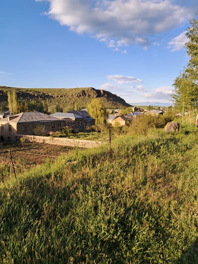 In the city of Artsvakar, Armenia, an image captures a rural landscape bathed in the golden light of late afternoon or early morning. The foreground is dominated by a dense field of tall, green grass, interspersed with patches of yellow wildflowers and darker, dried vegetation, casting long shadows. A low stone wall, constructed from irregular grey stones, stretches horizontally across the mid-ground, separating the grassy field from a cultivated plot of land that appears to be a garden or small farm, with rows of tilled soil and sparse green shoots.
Beyond the wall, a cluster of buildings forms a small village. The most prominent structure is a long, rectangular house with a grey, corrugated metal roof and beige walls. It features multiple windows and appears to be in moderate condition. Adjacent to it is another building with a similar roof but constructed with darker, rough-hewn stones. Several other smaller structures with varied rooflines are visible further in the distance, nestled among trees.
The background features a gently sloping hill covered in green grass and rocky outcrops. Tall, slender trees with sparse foliage are scattered throughout the scene, particularly on the left side near the hill and to the right of the village. The sky above is a vibrant blue, with scattered white and grey clouds, some of which are illuminated by the sun. In the far distance, a hazy line of mountains is visible on the horizon. The overall impression is one of a peaceful, somewhat rustic, and lived-in rural environment.