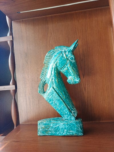 A teal horse head sculpture rests on a dark wooden shelf, positioned in front of a brown wooden cabinet or wall. The horse, painted with a textured brushstroke finish, is stylized with a geometric design, including a hole near its neck. Natural lighting creates a soft highlight on the wood surface where the statue stands. A shelf with turned spindles is visible on the left of the sculpture.