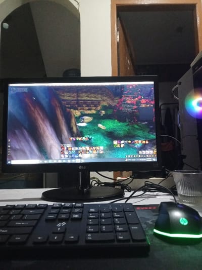 The scene is located in the city of Kpein, Liberia, depicting an active computer workstation in a dimly lit indoor environment. A black LG monitor displays a fantasy massively multiplayer online role-playing game (MMORPG), showing a vibrant green landscape with rocky structures and a detailed in-game user interface, including action bars, chat windows, and character information. In the foreground, a black full-size keyboard is positioned centrally, with an HP branded black optical mouse, illuminated by green LEDs, resting on a dark mousepad to its right. A black computer tower, emitting multi-colored RGB light, is partially visible on the far right. Numerous black cables are present on the light-colored desk surface, connecting the peripherals. To the right of the monitor, a clear, ribbed glass containing liquid is visible. The background features a light-colored wall with an arched opening on the left, where a faint, indistinct reflection of a human-like silhouette is discernible. An open wooden door frame on the right leads into a dark room. No discernible individuals are physically present in the immediate foreground.