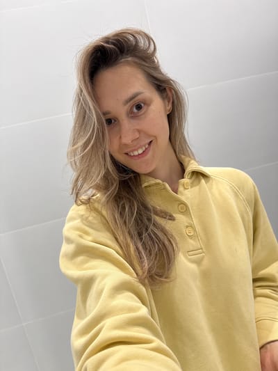 A headshot, likely a selfie, features a person, possibly female, with light blonde to light brown wavy hair. The person is looking directly into the camera with a gentle smile, their head tilted. They are wearing a light yellow long-sleeved shirt or sweatshirt with a collar and a placket featuring three matching yellow buttons. The background is a plain, light-colored surface, possibly a white or light gray wall with subtle grid lines indicating tiling. The lighting is bright and even, suggesting an indoor setting. No specific activity is depicted beyond posing for the photo. The person's expression appears friendly and pleasant. There is no visible text in the image. The location context provided is Новая Казмаска, Russia.