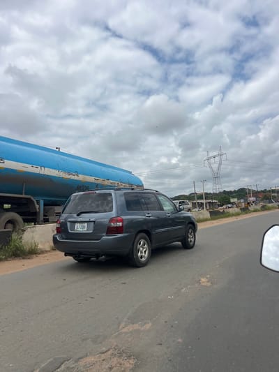 An overcast sky looms over a two-lane asphalt road. In the foreground, a dark gray SUV, possibly a Toyota Highlander, is driving on the right lane. Its license plate reads "BLF-20BH." To the left of the SUV, a large, blue tanker truck with white horizontal stripes is parked or moving slowly. The tanker truck appears to have some graffiti or markings on its side. The road is in moderate condition with some cracks and patches. In the background, there are buildings, shops, and vegetation, suggesting an urban or semi-urban environment. A tall electricity pylon with numerous wires is visible in the distance. The overall scene depicts a typical roadside view in Orifite, Nigeria, characterized by traffic, infrastructure, and a cloudy sky.