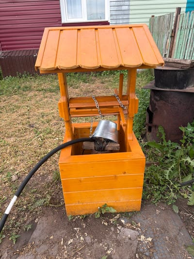In the rural village of Ilychimbatovo, Russia, a rustic scene unfolds. A bright orange wooden well, handcrafted and sturdy, stands proudly amidst overgrown grass and dirt. Its roof, also orange, shelters a metal bucket suspended by a chain, ready to draw water. A black hose with a white stripe curves from the left side of the frame, disappearing into the well's opening. To the right, a large, rusted metal barrel, possibly a stove, rests on a stand, its dark surface contrasting with the vibrant well. The background reveals a red-sided building with a white-trimmed window and a weathered green fence, further grounding the scene in its provincial setting. The ground is a mix of dry grass, soil, and patchy, gray ribbed material, hinting at the practical, utilitarian nature of the environment.