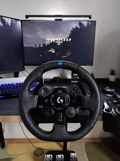 The scene depicts an indoor gaming setup located in Hrishteni, Bulgaria. A black Logitech G racing steering wheel, featuring a central "G" logo and a blue accent stripe at the 12 o'clock position, dominates the foreground. Below the steering wheel, a set of three metallic pedals rests on a cream-colored rug with irregular green patterns. The steering wheel is positioned in front of a light-colored wooden desk.
On the desk, two computer monitors are visible. The primary monitor, positioned centrally behind the steering wheel, displays a dark, forested road scene with the word "WEDNESDAY" in white text across the upper portion of the screen. A black LED light bar is mounted horizontally on top of this monitor. To the left, a secondary monitor is oriented vertically, displaying a dark, forested landscape matching the aesthetic of the primary screen.
Also on the desk, to the left, is a white mechanical keyboard with blue backlighting. To the right of the steering wheel, a black gaming mouse with blue accents, another Logitech peripheral (likely a shifter), and a small potted succulent plant are present. The desk surface is partially covered by a black mouse pad. The background consists of a light-colored, textured wall. No individuals are present.