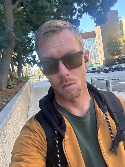 A light-skinned man wearing sunglasses, a dark green t-shirt, and a brown and black jacket with a black backpack stands outside on a sunny day. He has short blond hair and a light beard. Cars and buildings are visible in the background, along with trees and landscaping. The image is a selfie, taken at an angle. The man is facing the camera with a neutral expression.