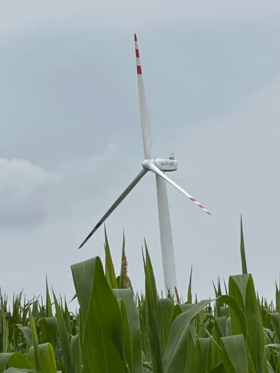 In Kolubaivtsi, Ukraine, a tall white wind turbine with red and white striped blades stands amidst a field of green corn stalks. The sky above is overcast and gray. The wind turbine has "SUZLON" written on its nacelle, the housing that contains the generator and gearbox. One of the turbine blades appears to be damaged or broken.