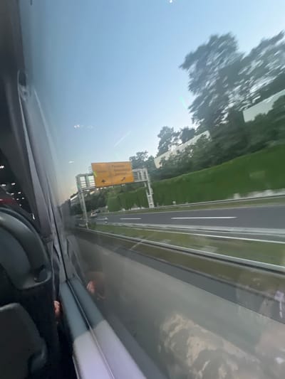 The image depicts a view from inside a vehicle, likely a bus, looking out the window. The primary focus is a yellow highway sign indicating directions to "Potsdam" and "Eisengrein". The sign is partially obscured by the window frame and reflections. The environment appears to be a highway in Kleinmachnow, Germany, with green barriers lining the side of the road and trees visible in the background. The sky is clear and blue, suggesting daytime. There are faint reflections on the window, including what appears to be interior seating and possibly some people. The overall scene captures a moment of travel on a highway.