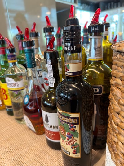 The image displays a collection of liqueur bottles, likely in a bar or retail setting, given the abundance of bottles and the presence of pour spouts on many of them. The bottles are arranged vertically, with some leaning against each other. They appear to be a variety of brands and colors, including clear, green, and dark liquids. A woven basket-like object is visible at the top of the image, partially obscuring some of the bottles. The lighting suggests an indoor environment, possibly with artificial light creating reflections on the glass bottles. The overall impression is one of a well-stocked bar or a display of alcoholic beverages. Some bottles have labels with visible text, such as "DRENNE", "DE CASSIS", "GORDON'S", and "ANDEMAN", indicating various types of liqueurs or spirits. The presence of pour spouts suggests the bottles are ready for use. The scene is static, with no people or action depicted. The focus is solely on the bottles themselves and their arrangement.
