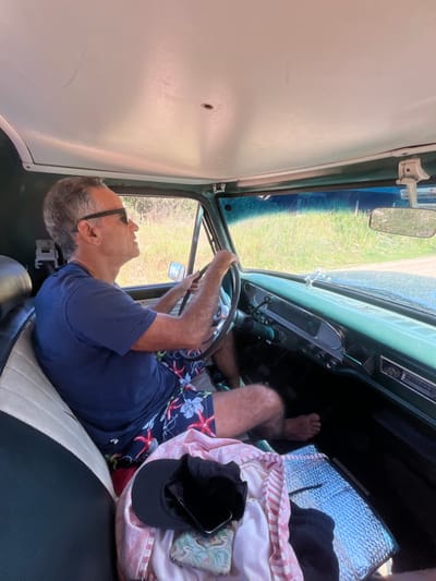 The image depicts the interior of an older model vehicle, located in La Barra, Uruguay, from the perspective of the front passenger seat. An adult male is operating the vehicle. He has short, graying hair, wears dark sunglasses, a dark blue t-shirt, and multi-colored swim trunks with red starfish and blue floral patterns. He is barefoot, with both hands on a dark steering wheel, gazing forward and slightly to his right through the windshield. The vehicle's interior features a light greenish-blue dashboard and door trim, an off-white headliner with a small central perforation, and a two-tone (cream and dark) driver's seat. A small, silver-toned winged ornament is affixed to the dashboard. On the front passenger seat, a black baseball cap covers a smartphone, alongside a light-colored patterned fabric and a small, green-patterned pouch. A silver reflective mat lies on the passenger side floorboard. Outside the vehicle, visible through the windows, is a bright, sunny environment dominated by tall green and yellowish grasses or brush, consistent with an unpaved road or natural area. A rearview mirror displays a white tag with dark text, which includes 'D.N.R.P.A.', hanging from its base.