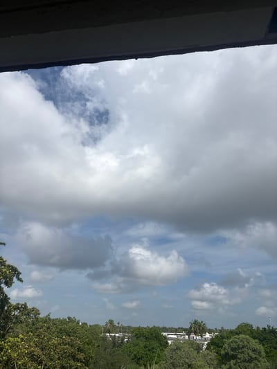 The image depicts a cloudy sky with some patches of blue visible. On the right side of the frame, there is a dense line of green trees, and beyond them, a few white buildings and palm trees can be seen. A dark structure, possibly a building or overhang, occupies the left side of the image. The lighting suggests it is daytime, likely late morning or early afternoon, given the brightness of the sky and the clouds. The weather appears to be mixed, with both bright and darker, more ominous clouds present. The scene is set outdoors, with the vegetation and buildings hinting at a tropical or subtropical coastal location. There are no people or explicit objects visible besides the natural elements and structures. No text is discernible in the image.