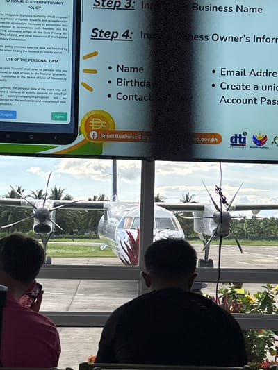 The image shows an indoor scene overlooking an airfield. Two people are looking at a phone. On the phone screen is an application for a National ID eVerify in the Philippines, requesting information such as business name, owner's information, name, birthday, contact, email address, and account password. Through a window, an airfield is visible, with several small aircraft parked on the runway. Palm trees and other vegetation can be seen in the background. The scene is located in Del Carmen, Philippines, according to reverse image search results of the image.