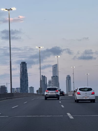 The image shows a highway with several cars, including a white car and a SUV, driving on the right lane. Streetlights line the side of the road. In the background, a city skyline features modern high-rise buildings. The sky is pale with some clouds. The image is taken at an angle.